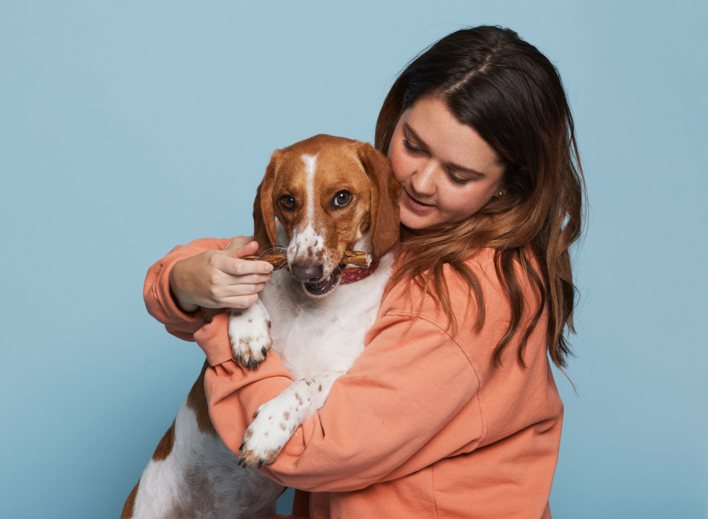 The girl is holding a dog that has a product from the Natural company in his mouth